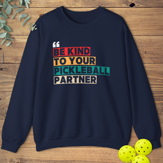 Navy blue sweatshirt with a stacked block design. White text reads 'BE KIND' on a red-orange block, 'TO YOUR' on a yellow-orange block, 'PICKLEBALL' on a teal-green block, and 'PARTNER' on a light grey-beige block. Small white quotation marks are at the top left