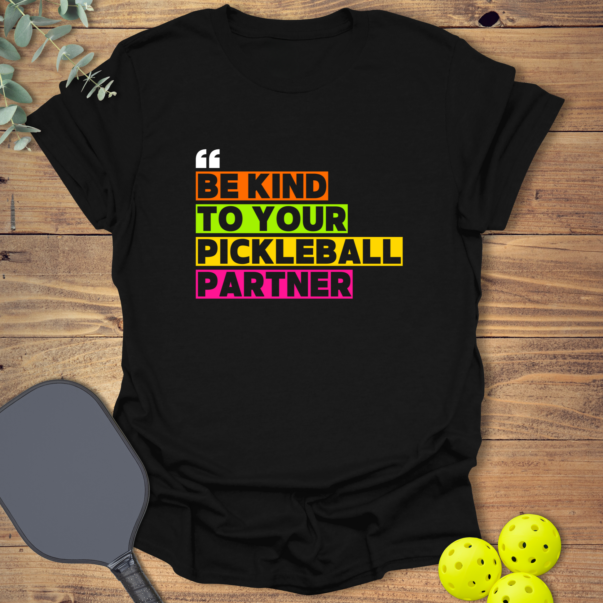 Black pickleball t-shirt with a stacked block design. White text reads 'BE KIND' on a red-orange block, 'TO YOUR' on a yellow-orange block, 'PICKLEBALL' on a teal-green block, and 'PARTNER' on a light grey-beige block. Small white quotation marks are at the top left