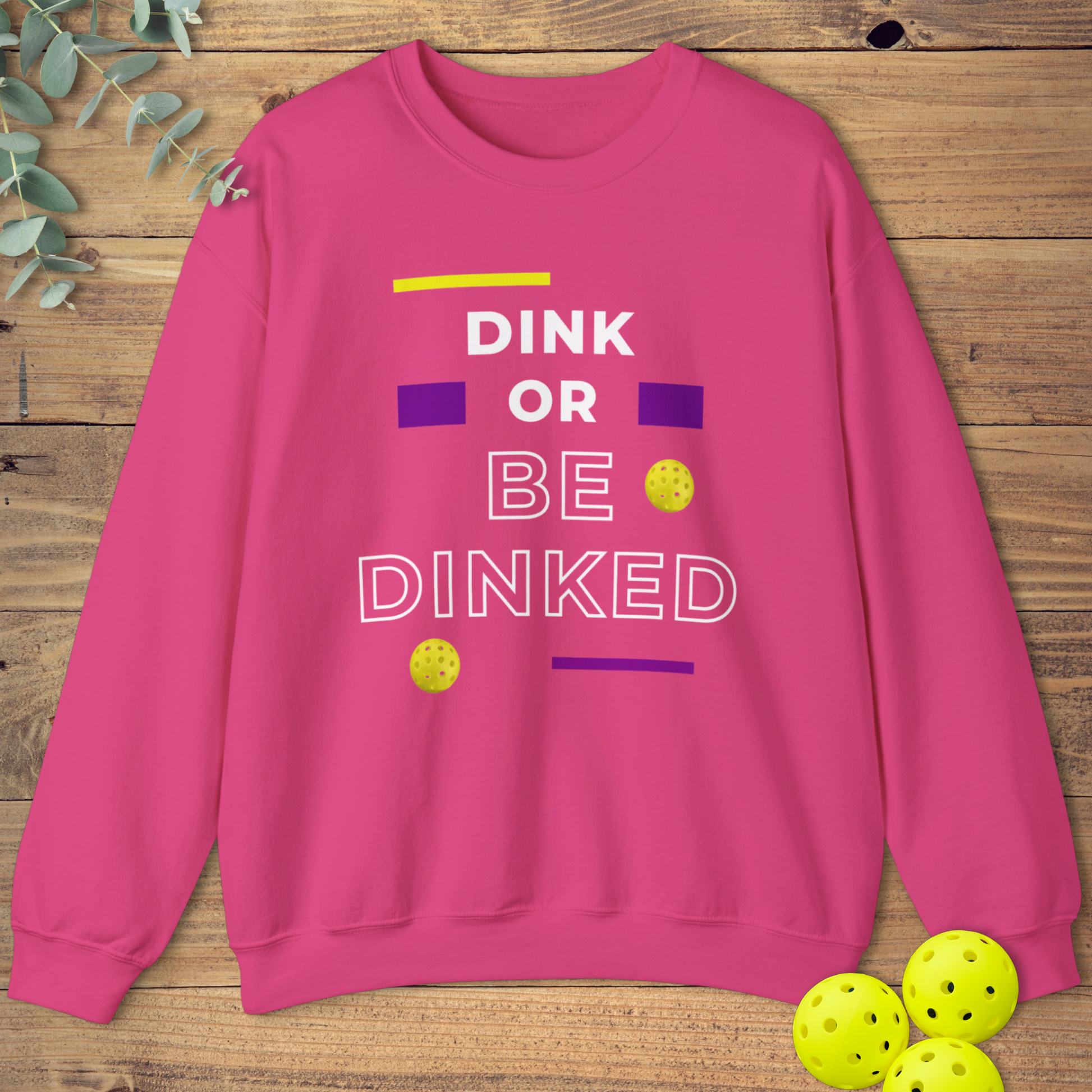 Pink pickleball sweatshirt with the phrase “Dink or Be Dinked” in bold white, purple, and yellow text, accented with small yellow pickleballs.