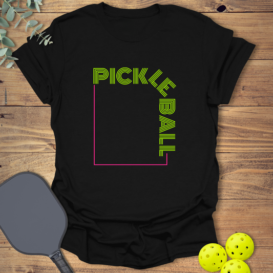 Black t-shirt with a modern geometric graphic. Neon green text spells 'PICKLEBALL' with 'PICKLE' at the top and 'BALL' rotated vertically on the right side. A pink outline forms the square frame