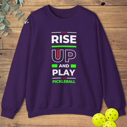 Purple sweatshirt with a stacked, motivational design. White text reads 'RISE', followed by large red outlined 'UP', 'AND', and 'PLAY'. 'PICKLEBALL' is smaller at the bottom. Red dots and yellow lines accent the text
