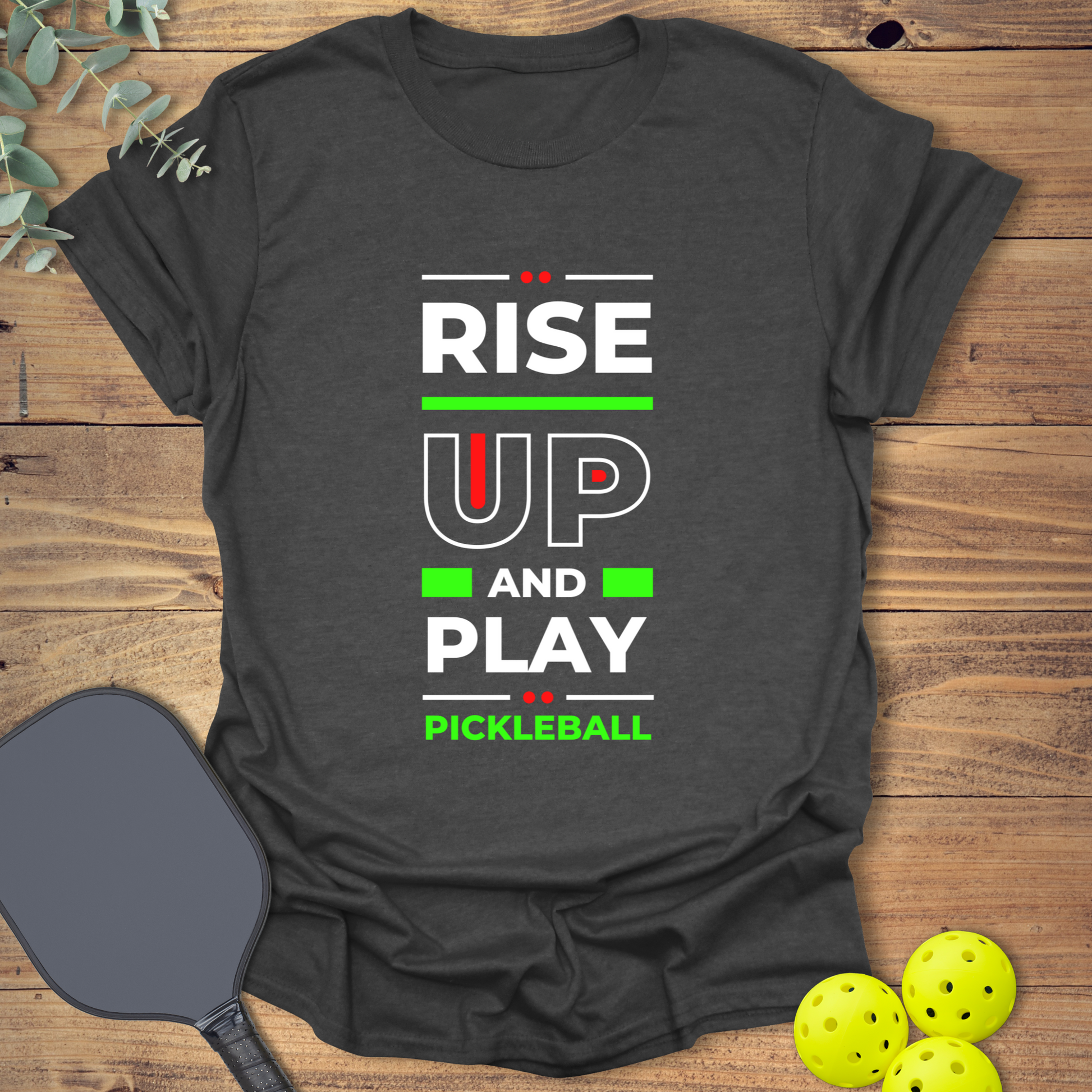 Heather grey t-shirt with a stacked, motivational design. White text reads 'RISE', followed by large red outlined 'UP', 'AND', and 'PLAY'. 'PICKLEBALL' is smaller at the bottom. Red dots and yellow lines accent the text.