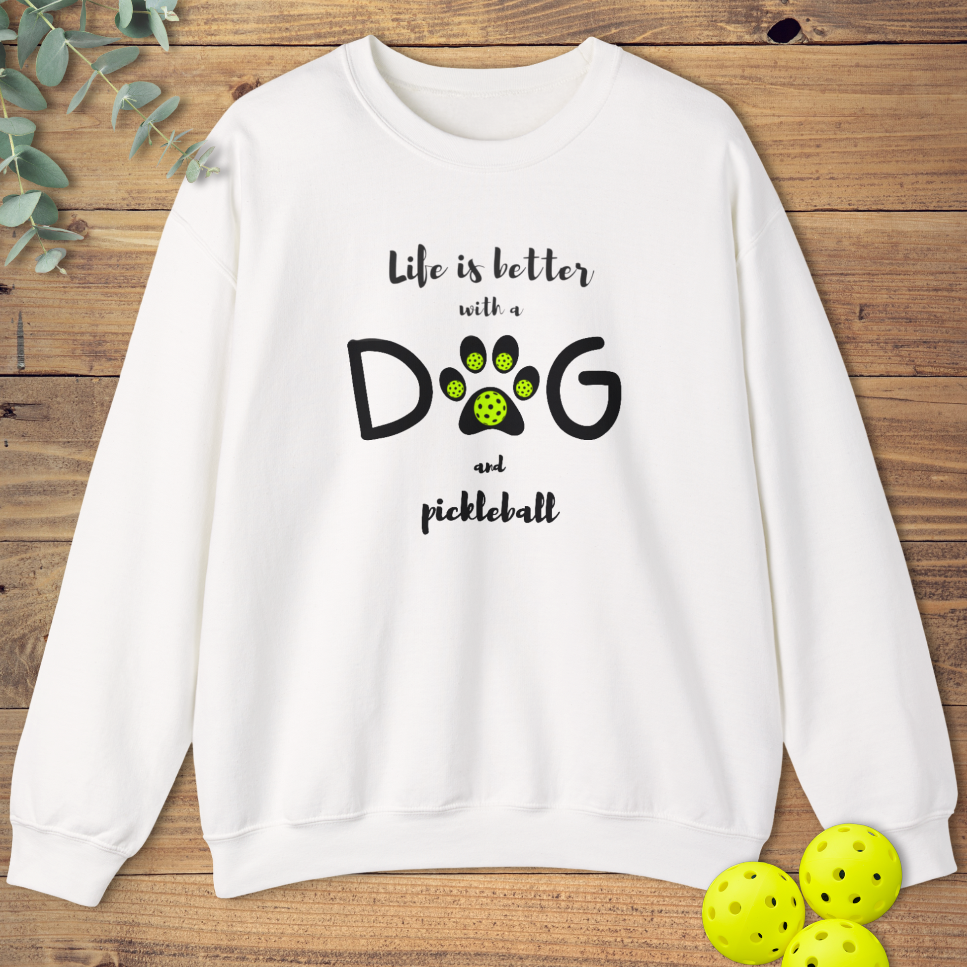 White graphic sweatshirt reading 'Life is better with a dog and pickleball'. The letter 'O' in 'DOG' is a black paw print with five neon green pickleball balls.