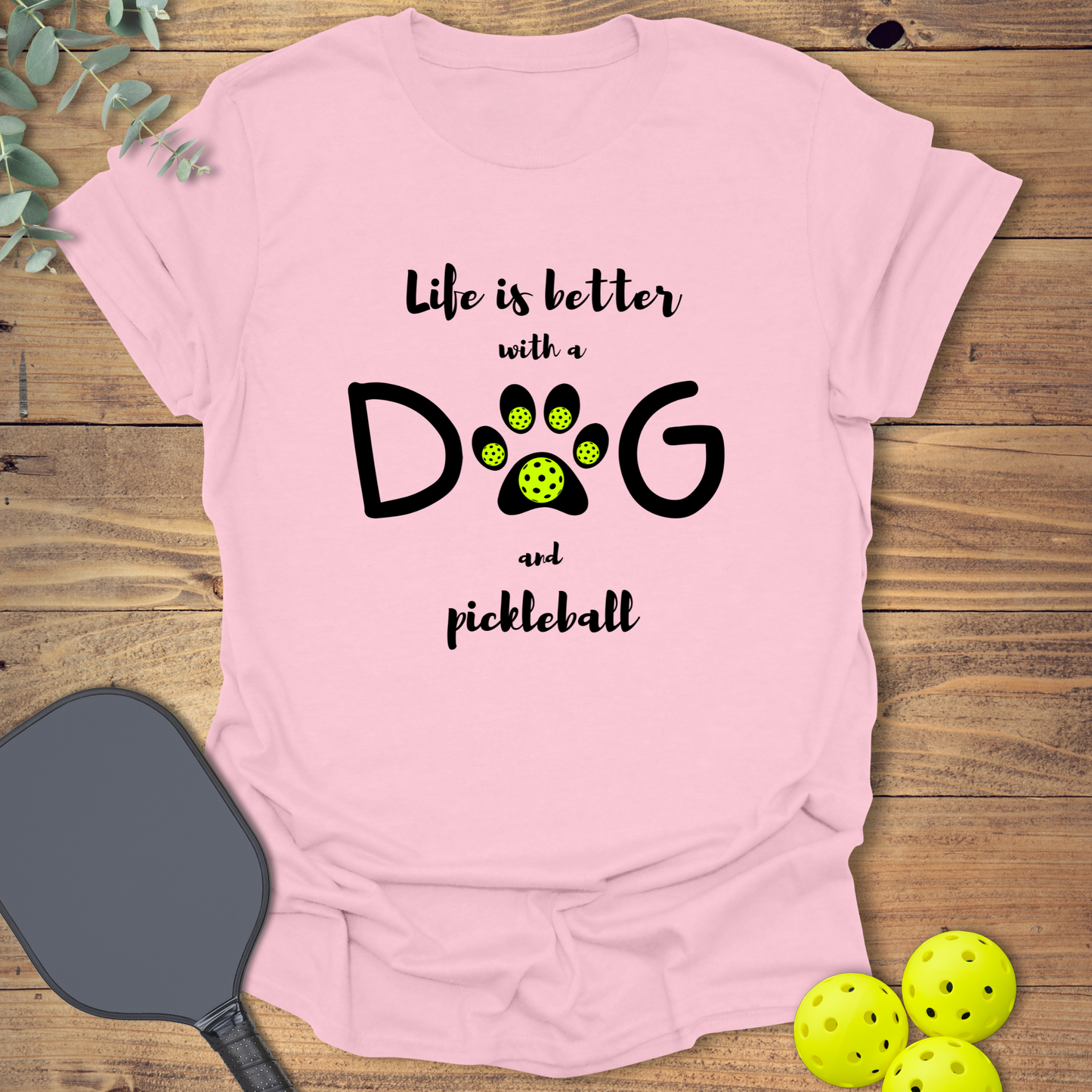 Pink graphic t-shirt reading 'Life is better with a dog and pickleball'. The letter 'O' in 'DOG' is a black paw print with five neon green pickleball balls.